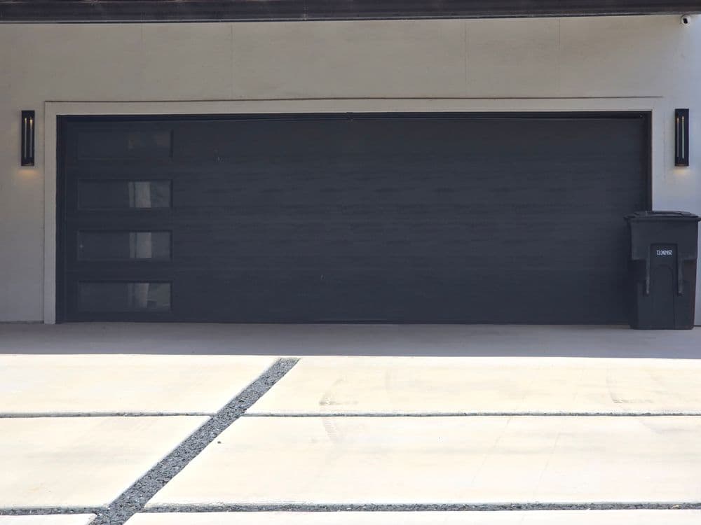 Modern black garage door with horizontal windows and a trash can beside it on a concrete driveway.