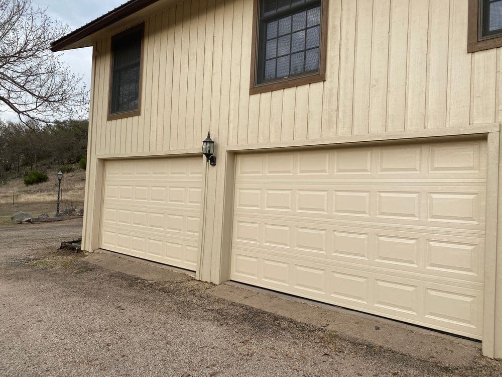 Two beige garage doors on a rustic yellow house exterior with a lamp post nearby.