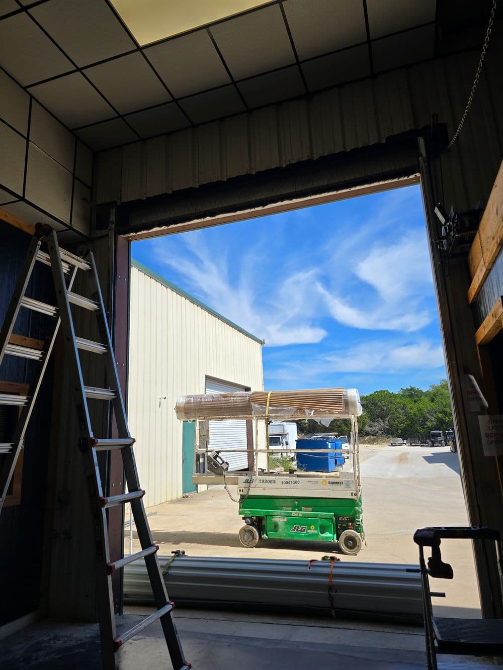 Interior view of a building with a ladder, scissor lift, and blue sky through an open door.