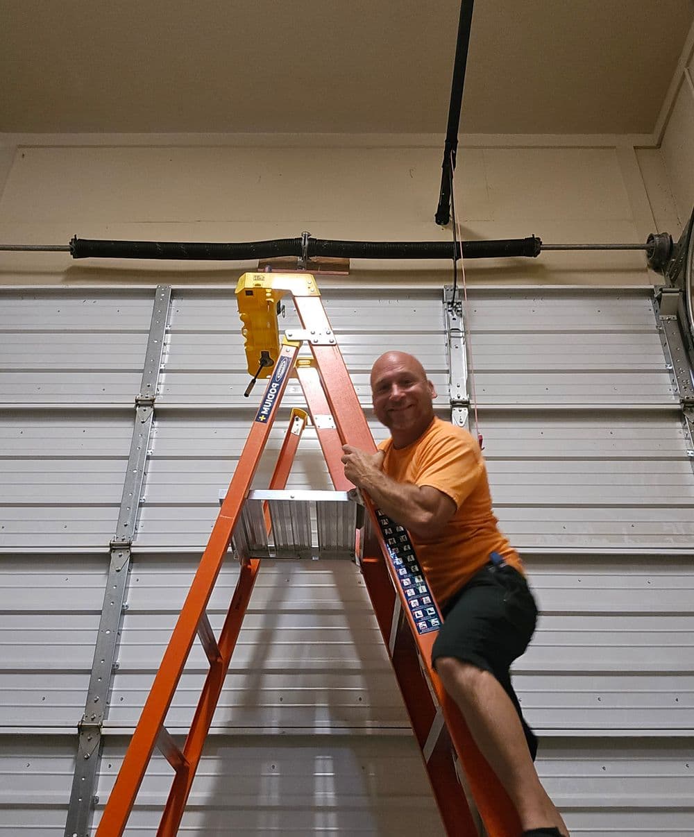 Man on orange ladder performing tasks in a garage with white walls and overhead equipment.
