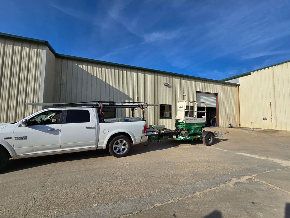White truck towing a green construction lift outside a warehouse under blue skies.