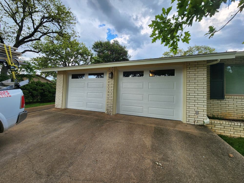 Two white garage doors on a brick house with a cloudy sky above and green trees nearby.
