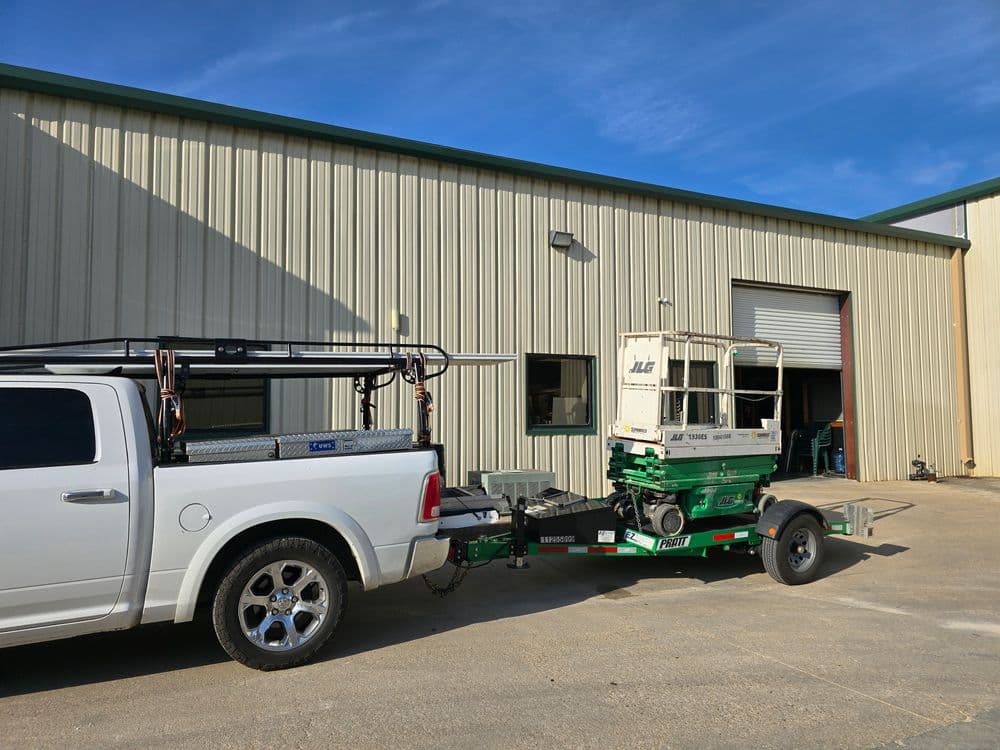 Pickup truck towing a scissor lift near a large metal building on a sunny day.