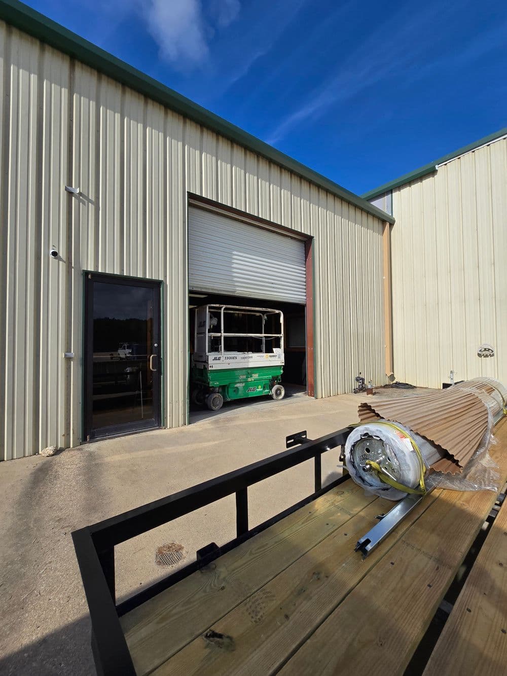 Exterior view of a warehouse with a green scissor lift and rolled-up material by the entrance.