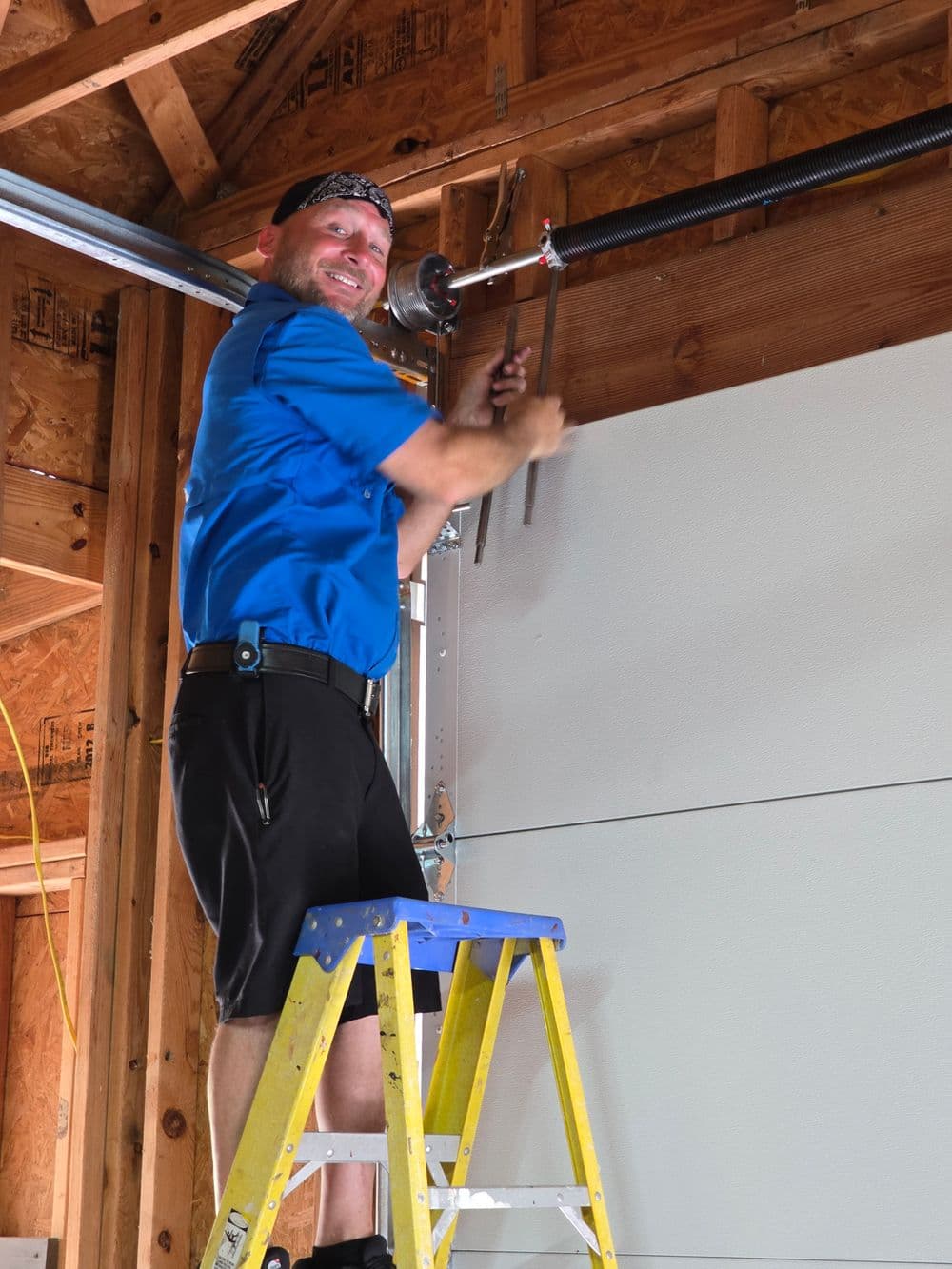 Technician in blue shirt repairs garage door mechanism on ladder with tools in hand.