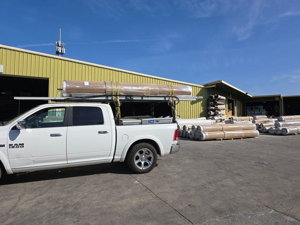 White pickup truck loaded with rolled materials outside a warehouse, sunny day, industrial area.