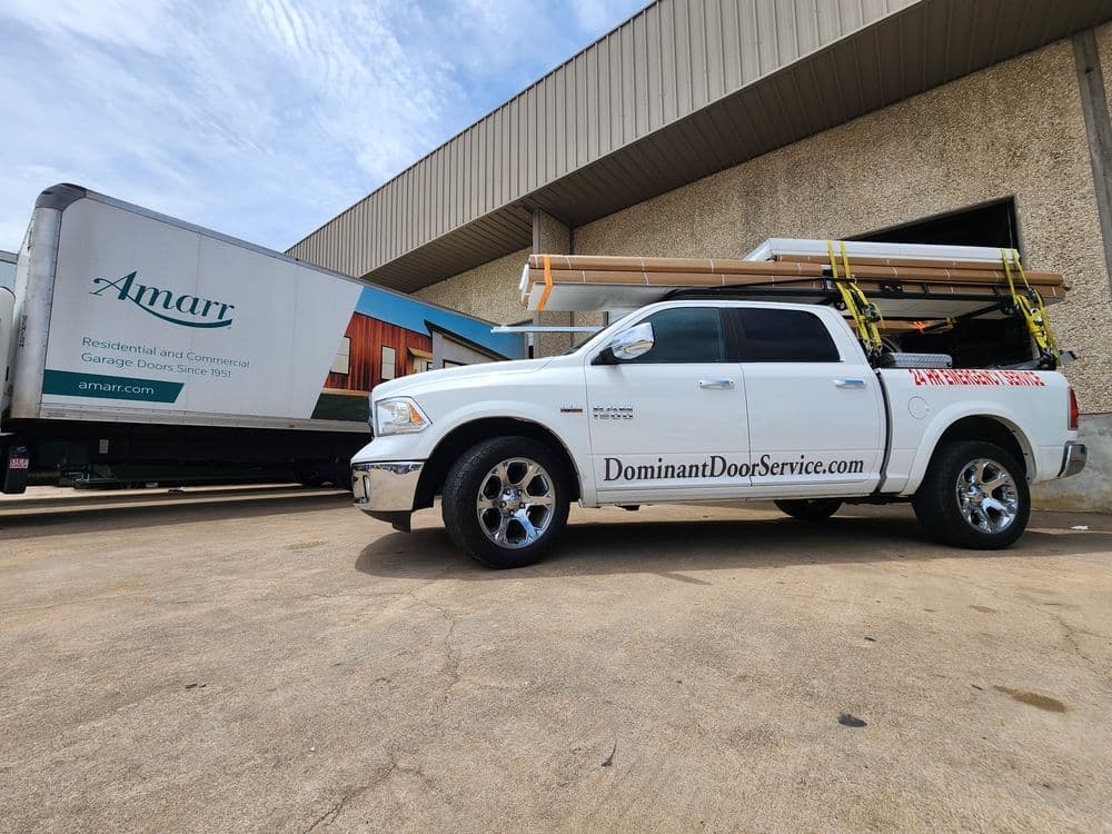 White service truck with "Dominant Door Service" branding parked beside an Amarr truck loaded with garage doors.