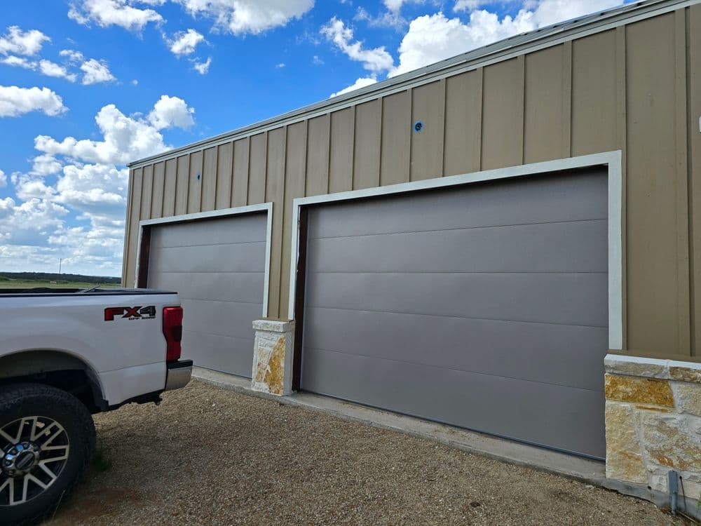 Modern two-car garage with sleek gray doors next to a pickup truck on gravel driveway.