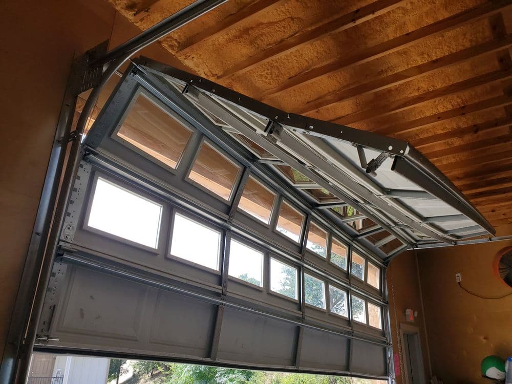 Modern garage door opened, revealing sunlight and outdoor view. Features large glass panels.