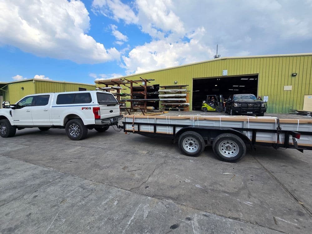 White truck with trailer loaded with lumber outside a warehouse under a blue sky.