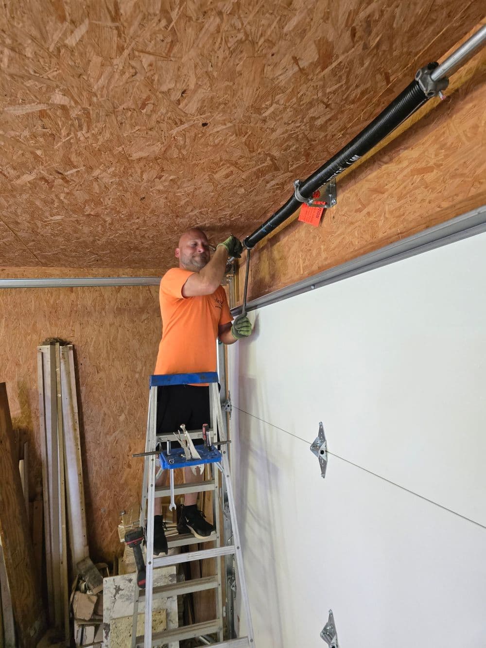 Man repairing garage door mechanism while standing on a ladder in a wooden garage.