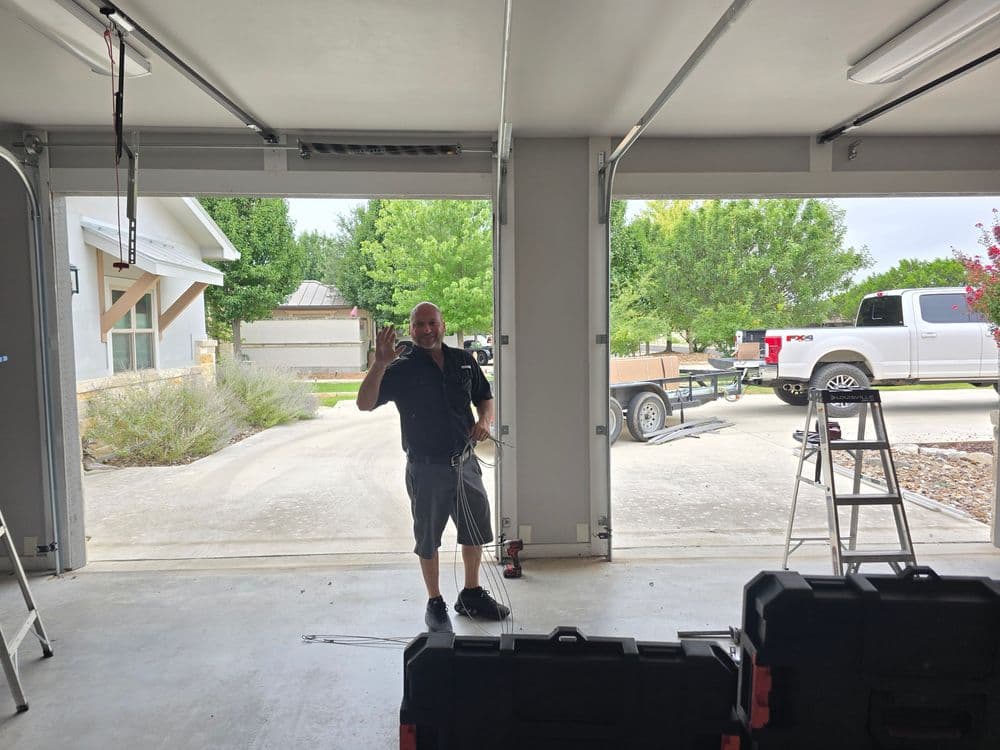 Man standing in a garage, waving, with a truck and trailer visible outside.