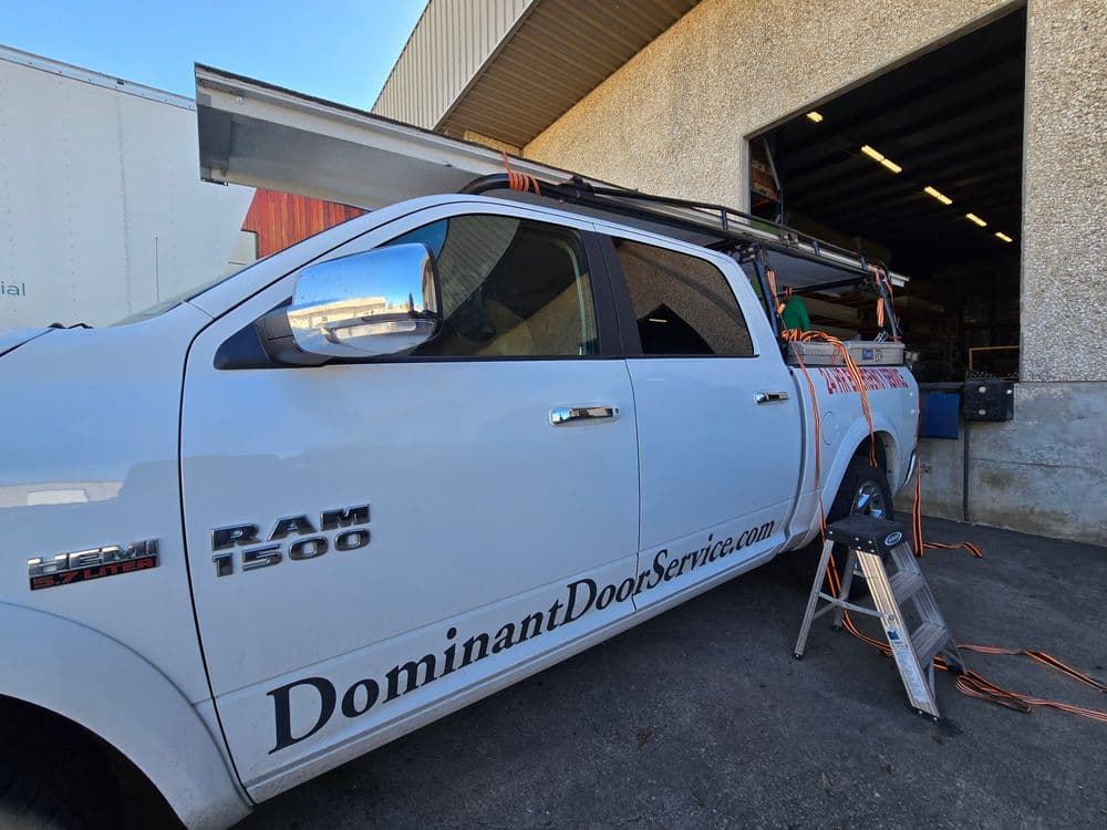 Dominant Door Service truck parked outside a warehouse with equipment and ladder visible.