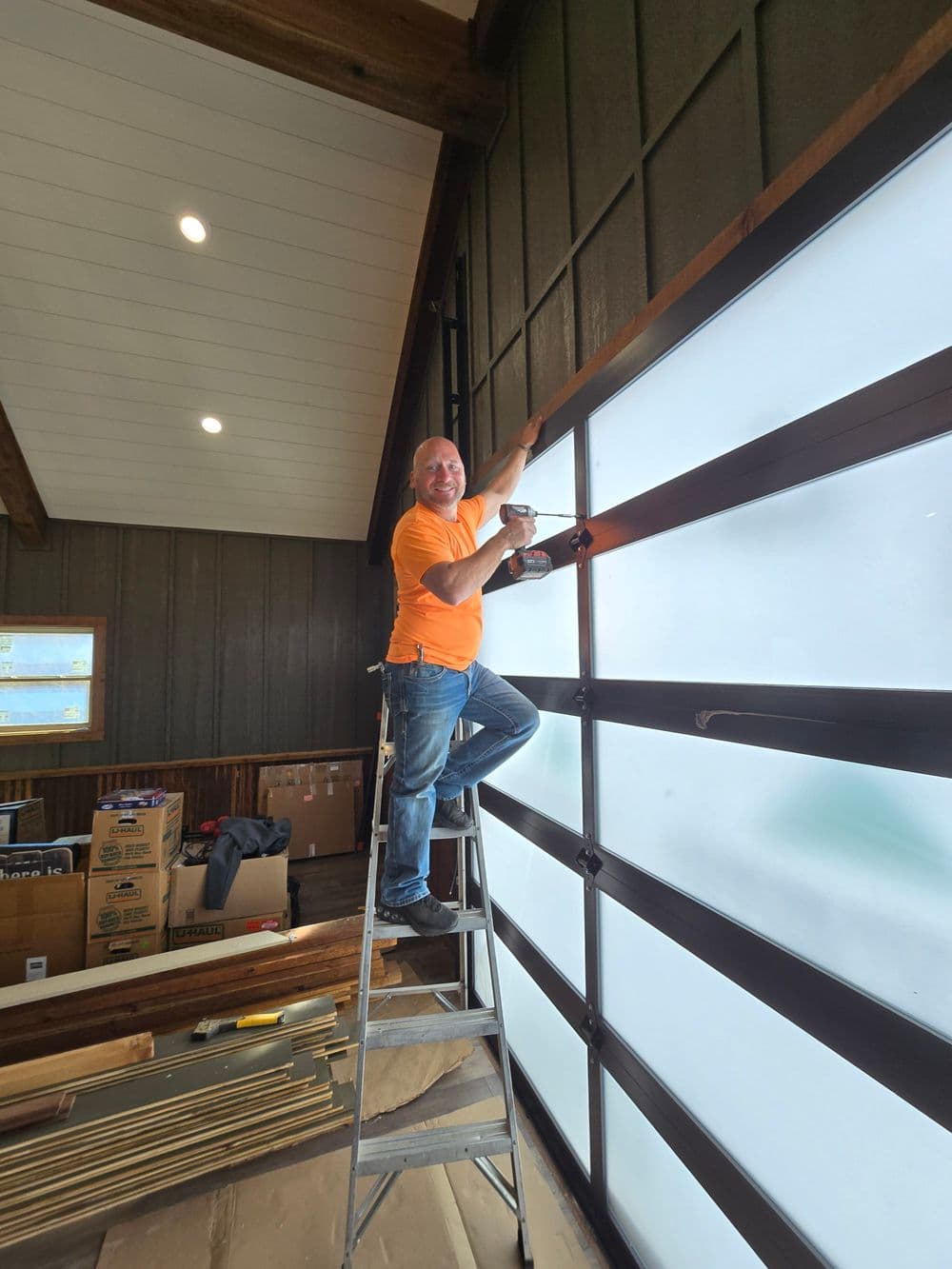 Man using a drill on a large glass garage door, standing on a ladder in a workshop.