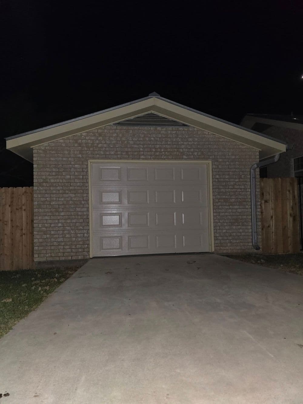 Brick garage with beige door and concrete driveway at night, surrounded by wooden fence.