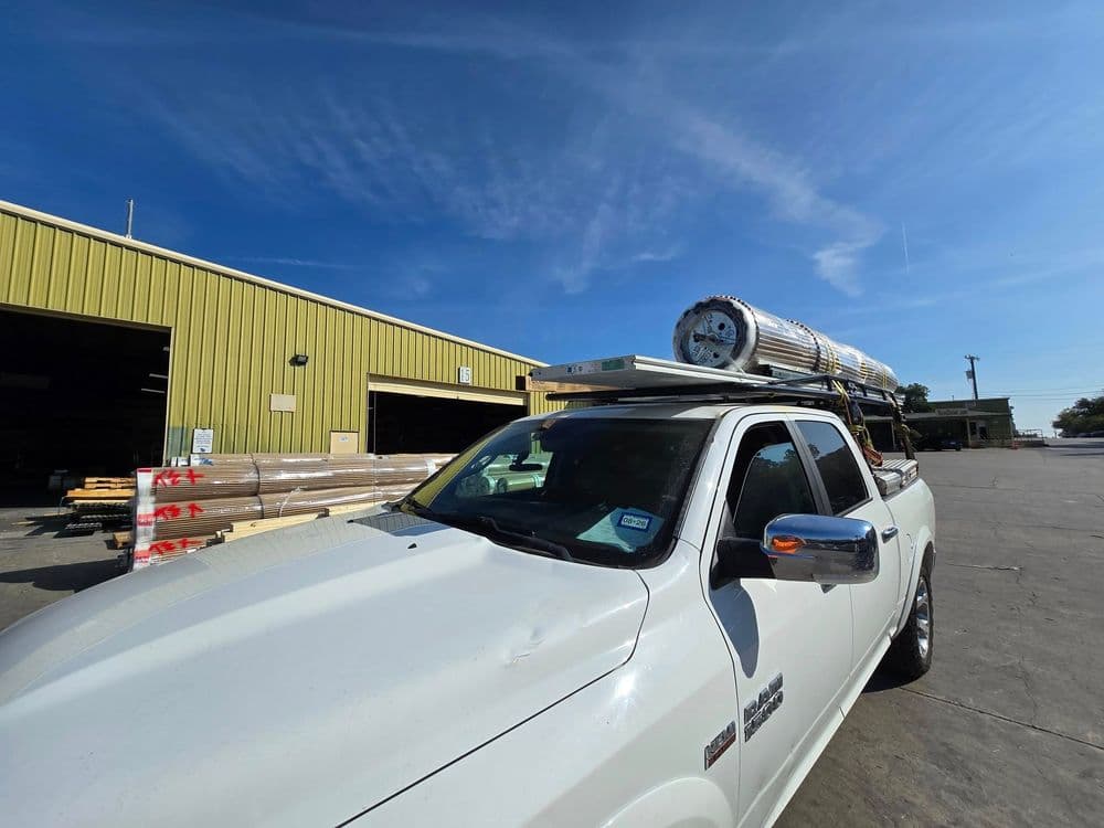 White pickup truck loaded with metal pipes parked outside a warehouse under clear blue sky.