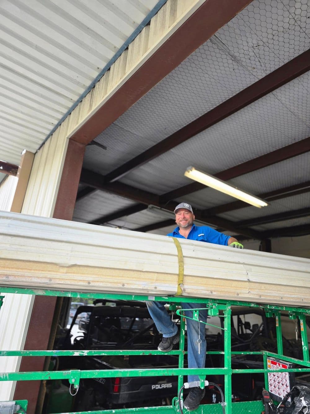Worker securing metal sheets on a green lift inside a warehouse with overhead lighting.