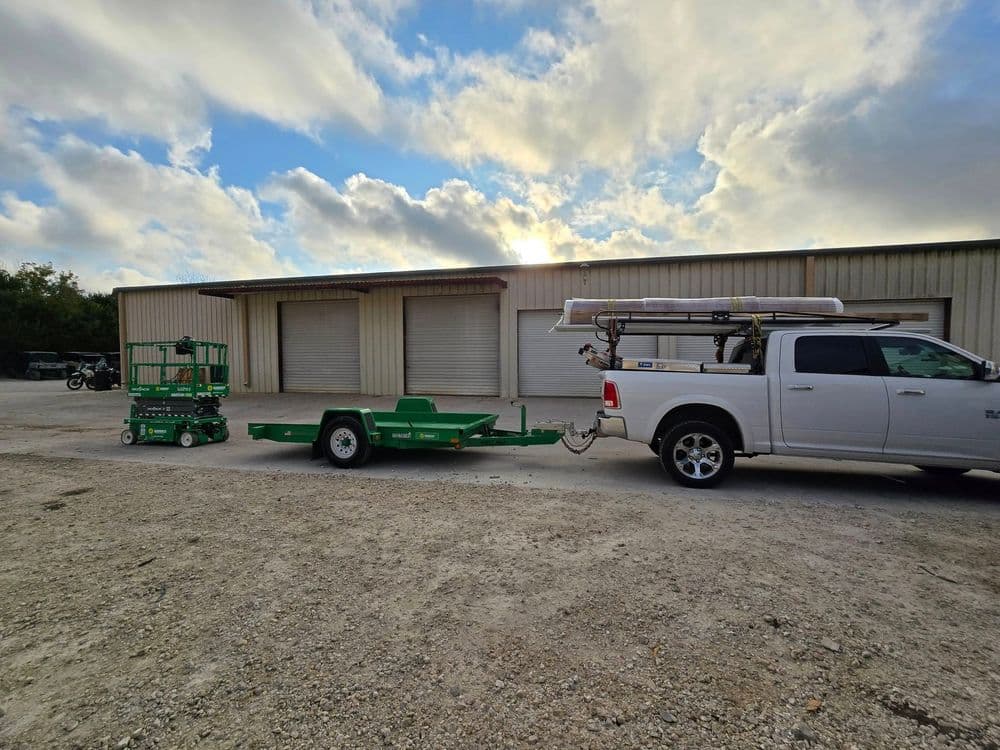 Pickup truck towing a green trailer with a scissor lift in front of industrial storage units.